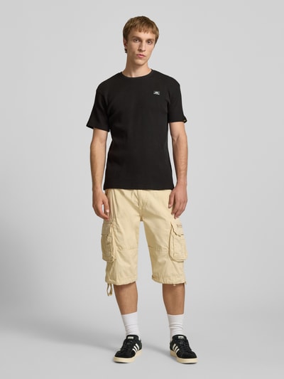 Männermodel trägt T-Shirt mit Label-Patch von Alpha Industries in Schwarz, Reine Baumwolle, Körpernahe Passform, Rundhalsausschnitt, Ganzkörperansicht von vorne. Rückenlänge bei Größe M: 66 cm