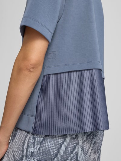 Kobieta nosi bluza 'Sweatshirt mit Rundhalsausschnitt' od Marc Cain, elastyczna mieszanka materiałów z dodatkiem wiskozy, prosty krój, okrągły dekolt, rękaw o dł. 1/2, widok z boku, detal tkaniny plisowanej.