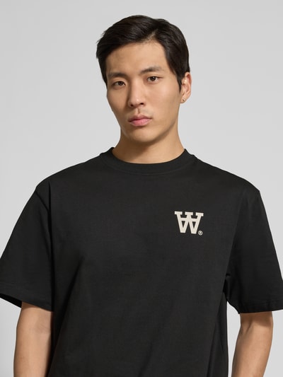 Męski model nosi T-Shirt mit Rundhalsausschnitt Modell 'Liam' od Wood Wood, czarny, bawełna, krój regular fit, label-print, zbliżenie na górną część ciała.