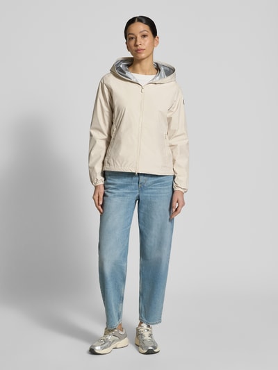 Frauenmodel trägt Regular Fit Jacke mit Zweiwegereißverschluss Modell 'Stella' von SAVE THE DUCK in Beige, hüftlang, glatte Textur, Ganzkörperansicht von vorne. Kontext: Damen Jacke, Eingrifftaschen mit Reißverschluss, Maße bei Größe S: Rückenlänge 60 cm, Schulterbreite 42 cm, Brustweite 52 cm, Ärmellänge 64 cm.