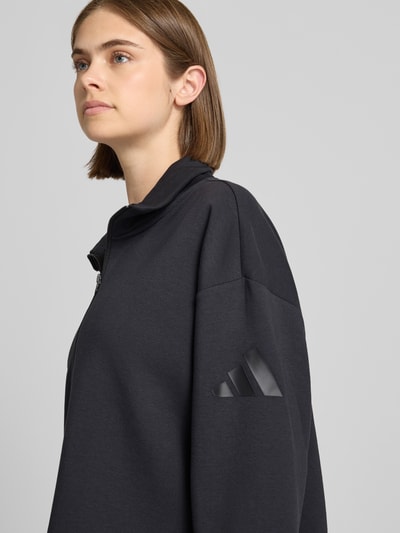 Vrouwenmodel draagt zwart Regular fit sweatshirt met katoen van adidas Sportswear, zilverkleurige ritssluiting, detailopname bovenlichaam.