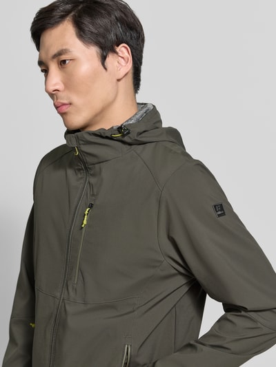 Męski model nosi Regular Fit Jacke mit mit Logo-Applikation von Killtec w kolorze khaki, kaptur z tunelem, wpuszczane kieszenie z zamkiem błyskawicznym, aplikacja z logo, długość tyłu w rozmiarze M: 72 cm, szerokość ramion w rozmiarze M: 45 cm, szerokość klatki piersiowej w rozmiarze M: 57 cm, długość rękawów w rozmiarze M: 67 cm, detaliczny zamek błyskawiczny, napoleońska kieszeń, zbliżenie na górną część ciała, widok z przodu.