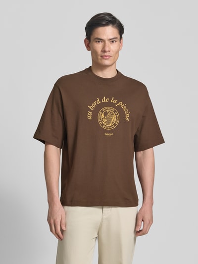 Männermodel trägt Relaxed Fit T-Shirt aus reiner Baumwolle Modell 'BOXYNORMAN GRAPHIC' von SELECTED HOMME in Braun mit gelbem Print, Rundhals, kurzarm, Jersey-Material, Ganzkörperansicht von vorne.