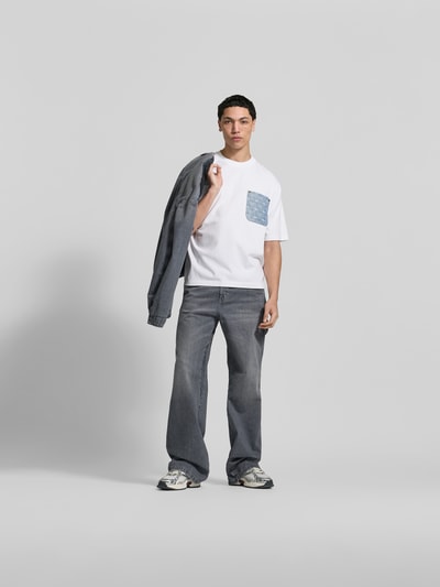 Männermodel trägt Loose Fit T-Shirt aus reiner Baumwolle von Dsquared2 in Weiß mit Denim-Brusttasche und Nieten, Ganzkörperansicht von vorne.