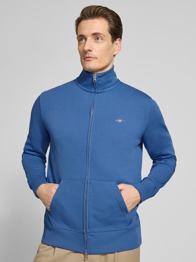 Męski model nosi Regular Fit Sweatjacke mit Stehkragen Modell 'Gield' od Gant, bawełniana mieszanka, z logo, zamek, kieszeń typu kangur, niebieski kolor, krój regular fit, widok całej sylwetki z przodu.