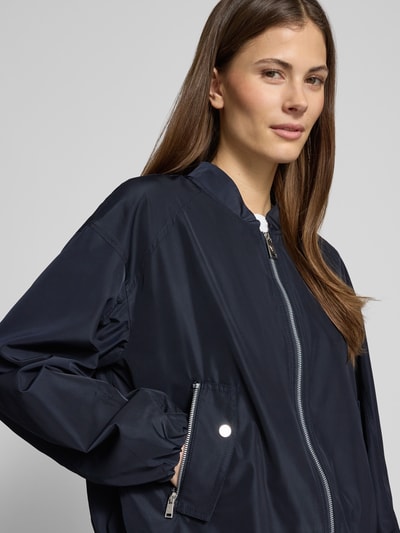 Kobiece modelka ma na sobie Jacke mit Reißverschlusstaschen od Fuchs Schmitt, krój regular fit, stójka, ciemnogranatowy kolor, tekstura gładka, zamek błyskawiczny, kieszenie na zamek, długość tyłu w rozmiarze 36: 52 cm, szerokość ramion w rozmiarze 36: 54 cm, szerokość w biuście w rozmiarze 36: 57 cm, długość rękawa w rozmiarze 36: 53 cm, zbliżenie na górną część ciała