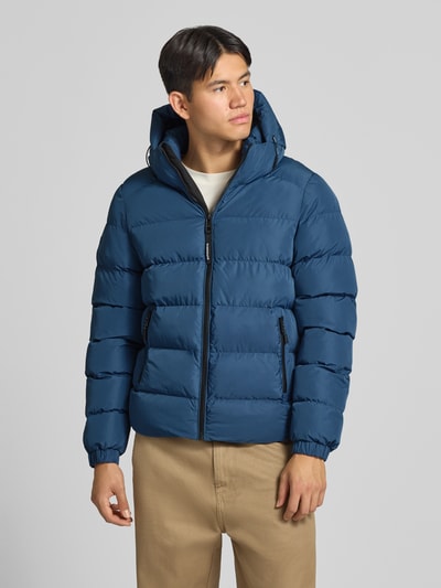 Männermodel trägt Steppjacke mit Label-Stitching und Kapuze von Superdry in Blau, sichtbare Reißverschlüsse, Seitentaschen, Ganzkörperansicht von vorne. Herren Steppjacke, gerader Schnitt, Kapuze mit Kordelzug, elastische Ärmelabschlüsse, Rückenlänge 67 cm, Schulterbreite 48 cm, Brustweite 58 cm, Ärmellänge 72 cm.