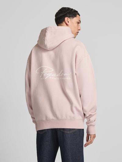 Männermodel trägt Oversized Hoodie mit Känguru-Tasche Modell 'Pike' von Pegador aus Baumwoll-Mix, Label-Print, Rückenlänge 71 cm, Ärmellänge 70 cm, Ganzkörperansicht Rückansicht.