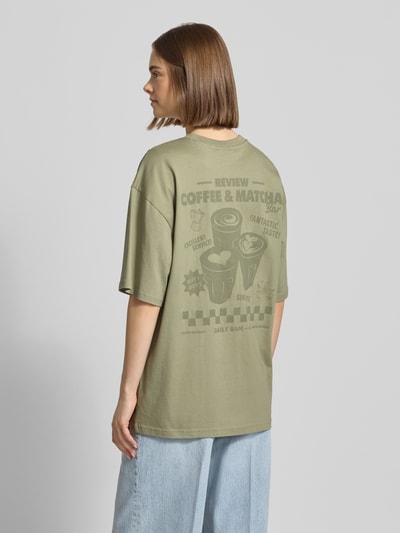 Modelka nosi Oversized T-Shirt mit Statement-Print von REVIEW, bawełna, krój oversize, okrągły dekolt, nadruk, długość tyłu w rozm. S: 70 cm, widok całej sylwetki z tyłu