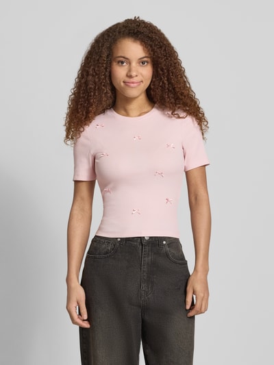 Kobiece model prezentuje jasnoróżowy Slim Fit T-Shirt aus Baumwoll-Mix Modell 'FEMKE' od Only, gładki, z kokardkami, okrągły dekolt, krótkie rękawy, drobny prążek; widok na cały przód górnej części ciała.