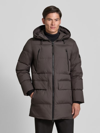 Männermodel trägt Regular Fit Daunenjacke mit Kapuze von Marc O'Polo in Dunkelbraun mit Zweiwege-Reißverschluss und Pattentaschen, Ganzkörperansicht von vorne.