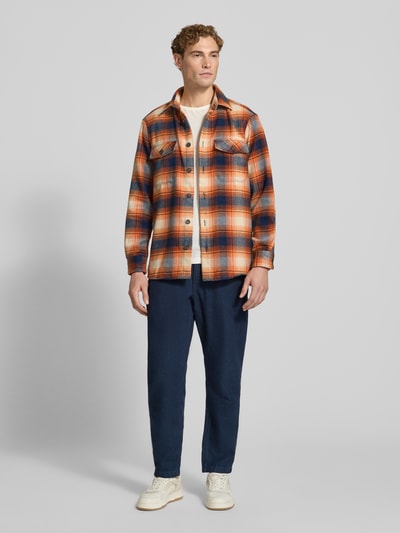 Männermodel trägt Regular Fit Hemdjacke mit Brusttaschen von Christian Berg Men, kariert in Orange, Blau und Weiß, Ganzkörperansicht von vorne. Kentkragen, durchgehende Knopfleiste, markante Brusttaschen, glatte Textur, Rückenlänge 71 cm, Schulterbreite 43 cm, Ärmellänge 68 cm.