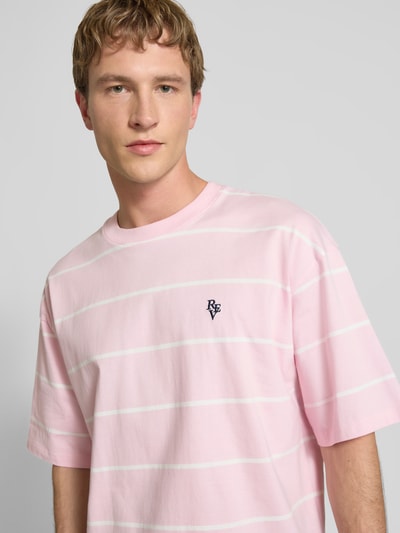 Männermodel trägt T-Shirt mit Rundhalsausschnitt und Logo-Stitching von REVIEW in Rosa mit weißen Streifen, reine Baumwolle, Loose Fit, Nahaufnahme Oberkörper.