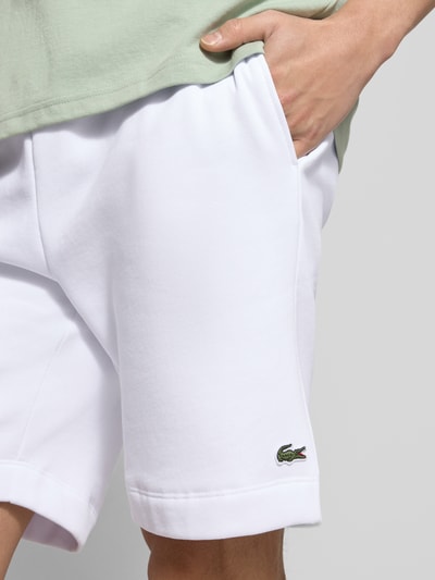 Męski model nosi Regular Fit Sweatshorts aus Baumwoll-Mix od Lacoste, biała jednolita struktura, detal z logo, krój regular fit, bawełniany materiał typu sweat, widok z przodu, zbliżenie na górną część nóg.