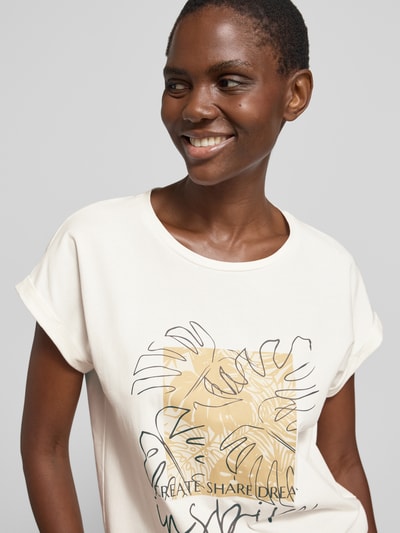 Frauenmodel trägt T-Shirt mit fixiertem Ärmelaufschlag Modell 'RINA' von Soyaconcept in Creme mit grafischem Motiv-Print, Nahaufnahme Oberkörper. Baumwoll-Mix mit Stretch-Anteil, gerader Schnitt und Rundhalsausschnitt erkennbar.