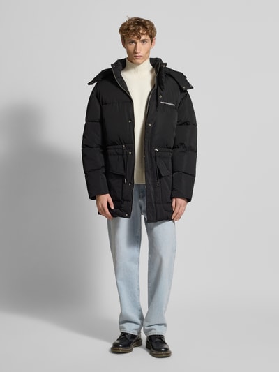 Männermodel trägt schwarzen Parka mit Kapuze Modell 'Briska' von Don't Waste Culture, lockerer Schnitt, Ganzkörperansicht von vorne.