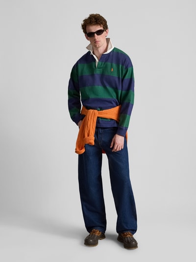 Männermodel trägt Oversized Pullover aus reiner Baumwolle Modell '6/1' von Polo Ralph Lauren, breite grün-blaue Streifen, dunkle Jeans, orangefarbener Pullover um die Hüfte, neutrale Texturen, Herren Pullover von Polo Ralph Lauren, Ganzkörperansicht von vorne.