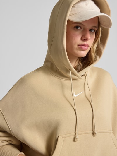 Frauenmodel trägt Oversized Hoodie mit Logo von Nike in Beige aus Baumwoll-Mix mit Känguru-Tasche, Nahaufnahme Oberkörper.