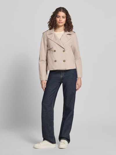 Modelka kobieca nosi Regular Fit Trenchcoat mit Umlegekragen od Review, jasny beż, czysta bawełna, dwurzędowe guziki, kieszenie, długość pleców w rozmiarze S: 55 cm, szerokość ramion w rozmiarze S: 45 cm, szerokość klatki piersiowej w rozmiarze S: 53 cm, długość rękawa w rozmiarze S: 62 cm, pełny widok z przodu