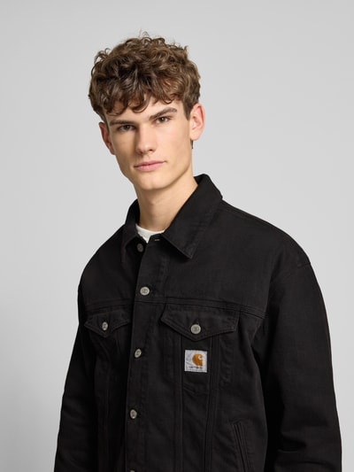 Männermodel trägt schwarze Jeansjacke mit Label-Detail Modell 'EVERSON' von Carhartt Work In Progress, Nahaufnahme Oberkörper.