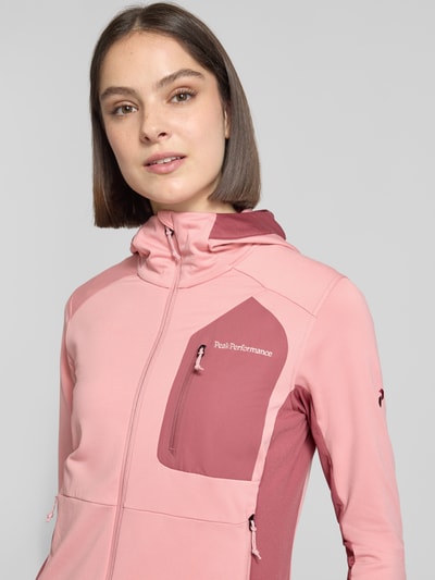 Frauenmodel trägt Jacke mit Logo-Stitching von PEAK PERFORMANCE in Rosa mit Elastischer-Material-Mix, körpernahe Passform, Kapuze, Reißverschluss und Brusttasche mit Reißverschluss, Nahaufnahme Oberkörper.
