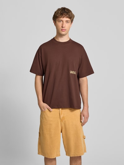 Mężczyznę model nosi t-shirt mit Label-Stitching und Rundhalsausschnitt von ANOTHER COTTON LAB, bawełna, krój relaxed fit, długość tyłu w rozmiarze M 69 cm, brązowy kolor, gładka struktura. Ganzkörperansicht von vorne.