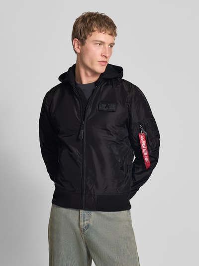 Männermodel trägt Herren Blouson mit Logo-Anhänger Modell 'MA-1 TT' von Alpha Industries, schwarze Farbe, Ganzkörperansicht von vorne, lockere Passform, Kapuze aus Sweat, durchgehender Reißverschluss, Armtasche mit Logo-Anhänger, Label-Patch, elastische Abschlüsse, Rückenlänge bei Größe M 69 cm, Schulterbreite 46 cm, Brustweite 54 cm, Ärmellänge 67 cm.