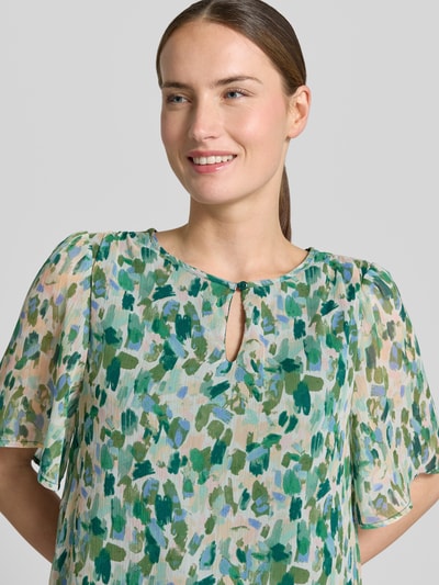 Modelka kobieca nosi Blusenshirt mit Schlüsselloch-Ausschnitt Modell 'EMMA' od Vila, krój regular fit, wzór allover, długość tyłu 60 cm, długość rękawa 29 cm, pastelowe zielone i niebieskie plamy, zwiewna tekstura, zbliżenie na górną część ciała z przodu.