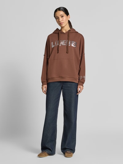 Frauenmodel trägt Oversized Hoodie aus reiner Baumwolle Modell 'L(I)EBE' von miss goodlife in Braun mit Pailletten, Känguru-Tasche, gerippten Abschlüssen, Ganzkörperansicht von vorne.
