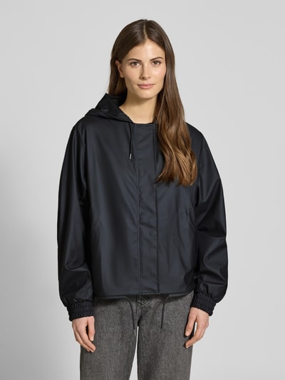 Frauenmodel trägt Jacke mit Abdeckleiste von Rains in Schwarz, gerade Passform, Kapuze, Reißverschluss, elastische Ärmelabschlüsse, Ganzkörperansicht von vorne. Rückenlänge bei Größe S: 58 cm, Schulterbreite: 43 cm, Brustweite: 55 cm, Ärmellänge: 63 cm, Tunnelzug im Saum.
