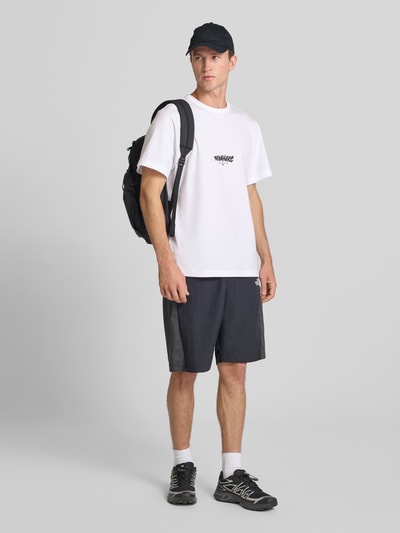 Männermodel trägt weißes Relaxed Fit T-Shirt aus reiner Baumwolle mit Logo-Print von The North Face, schwarze Shorts, Ganzkörperansicht von vorne.