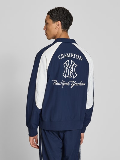 Männermodel trägt Relaxed Fit Tracksuit mit Logo-Patch von Champion in Dunkelblau und Weiß, Stehkragen, Rückenansicht, Kontext Herren Tracksuit, elastische Abschlüsse, Reißverschluss, Bundweite 39 cm, Rückenlänge 68 cm, Schulterbreite 42 cm, Ärmellänge 49 cm, Innenbeinlänge 72 cm. Ganzkörperansicht Rückansicht.