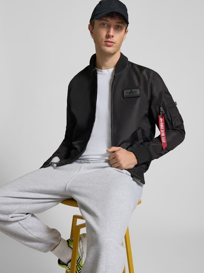 Männermodel trägt schwarzen Blouson mit Logo-Anhänger Modell 'MA-1 TT' von Alpha Industries, lockere Passform, Ganzkörperansicht von vorne