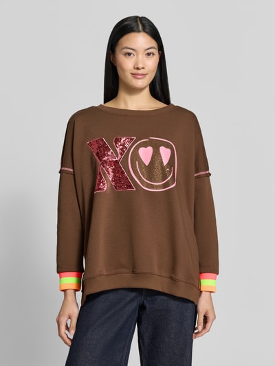Frauenmodel trägt Oversized Sweatshirt aus reiner Baumwolle Modell 'XO Heartface' von miss goodlife, braun mit Statement-Print, Rundhalsausschnitt, Ziernähte, elastische Abschlüsse, kräftige Neonfarben an Ärmeln, Ganzkörperansicht von vorne.
