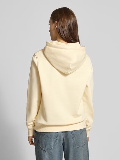 Kobiece model prezentuje Regular Fit Hoodie aus Baumwoll-Mix od Tommy Jeans, jednolity kremowy kolor, bluza z kapturem, materiał: bawełniany mix, detal: nadruk z logo, długie rękawy, ściągacze, zdjęcie całej sylwetki z tyłu.
