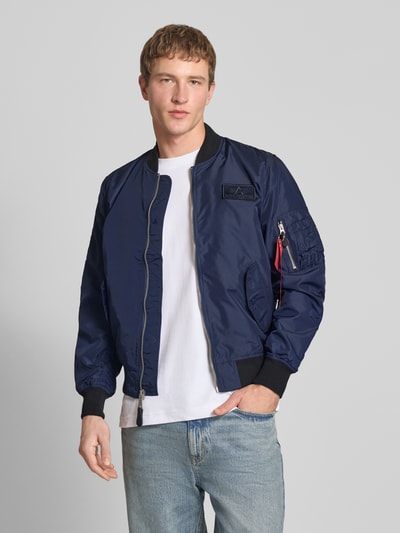 Męski model nosi Regular Fit Bomberjacke mit Logo-Patch od Alpha Industries, kolor granatowy, krój regular fit, stójka, kieszenie, zamek, naszywka logo, kieszeń na rękawie, długość pleców w rozmiarze M: 65 cm, szerokość ramion w rozmiarze M: 45 cm, szerokość w rozmiarze M: 54 cm, długość rękawa w rozmiarze M: 69 cm, całkowity widok z przodu.