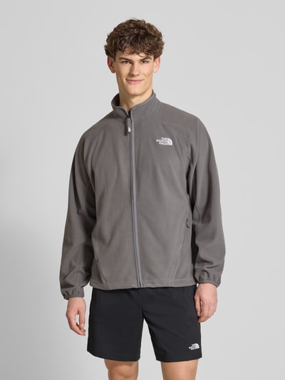Männermodel trägt graue Fleecejacke mit Label-Stitching von The North Face, Stehkragen, Reißverschluss, Regular Fit; Ganzkörperansicht von vorne.