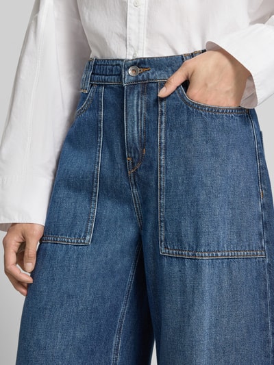 Frauenmodel trägt Wide Fit Jeans mit Baumwoll-Anteil Modell 'Derive' von DRYKORN in Blau mit aufgesetzten Taschen, Nahaufnahme Oberkörper. Material-Mix, Knopf- und Reißverschluss, Bundweite 40 cm, Innenbeinlänge 67 cm.