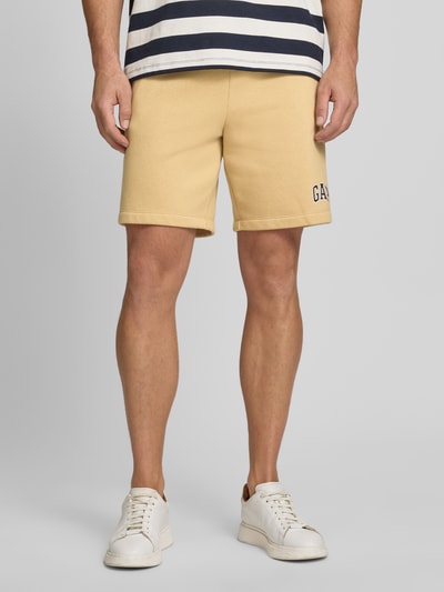 Männermodel trägt Sweatshorts mit Logo und elastischem Bund von GAP aus Baumwoll-Mix, Regular-Fit, Eingrifftaschen, Bundweite 40 cm, Innenbeinlänge 21 cm, sandfarbene Optik, Ganzkörperansicht von vorne.