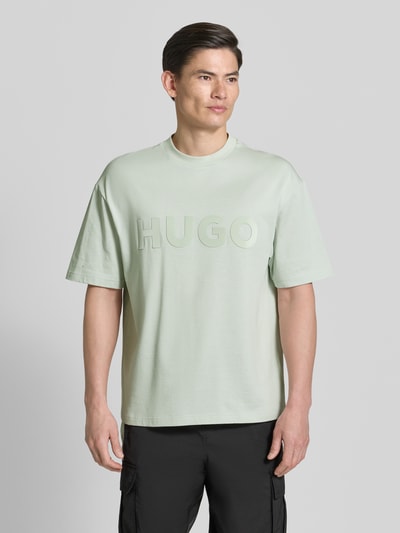 Männermodel trägt Relaxed Fit T-Shirt aus reiner Baumwolle Modell 'DINKEE' von HUGO in Optik: Unifarben mit 3D Label-Print, Rundhals-Ausschnitt, Ärmelform: Kurzarm, Materialverarbeitung: Jersey, Nahaufnahme Oberkörper.
