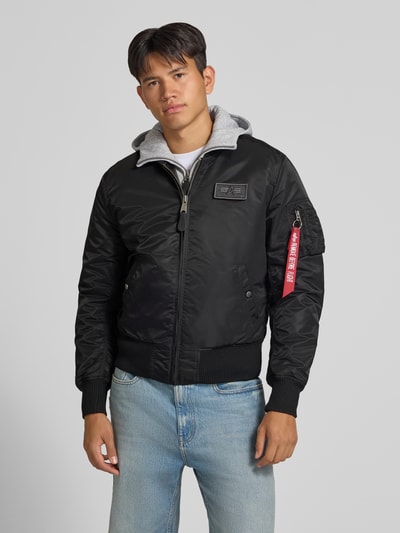 Männermodel trägt Jacke mit Reißverschlusstasche am Ärmel von Alpha Industries in Schwarz mit grauem Kapuzeinsatz, Nahaufnahme Oberkörper. Regular Fit, gerippter Stehkragen, Label-Patch, Eingrifftaschen mit Druckknopfverschluss, Brustweite 54 cm, Ärmellänge 72 cm.
