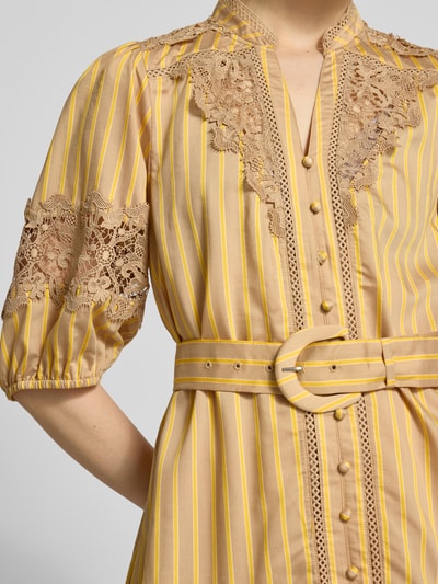 Frauenmodel trägt Knielanges Kleid mit Spitze und Gürtel Modell 'CATLEYA' von SUNCOO PARIS aus Viskose-Mix, Taillierter Schnitt, Stehkragen, V-Ausschnitt, Gürtel, beige mit gelben Streifen und Spitzen-Details, Nahaufnahme Oberkörper.