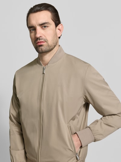 Męski model nosi bomberkę z prostym krojem marki CK Calvin Klein, model 'MATTE', beżowa, kieszenie zapinane na zamek błyskawiczny, stójka, elastyczne wykończenia, zdjęcie: zbliżenie na górną część ciała z przodu, długość tyłu w rozmiarze M: 67 cm, szerokość ramion w rozmiarze M: 47 cm, szerokość klatki piersiowej w rozmiarze M: 58 cm, długość rękawów w rozmiarze M: 68 cm