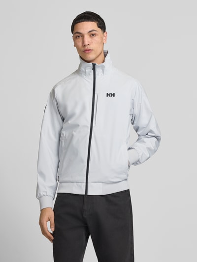 Männermodel trägt Bomberjacke mit Stehkragen Modell 'RACING' von Helly Hansen, gerader-Schnitt, Logo-Print, seitliche Eingrifftaschen, Reißverschluss, gerippte-Abschlüsse, hellgraue Farbe, glatte Struktur, Ganzkörperansicht von vorne. Rückenlänge 68 cm, Schulterbreite 43 cm, Brustweite 58 cm, Ärmellänge 68 cm, Größe M.