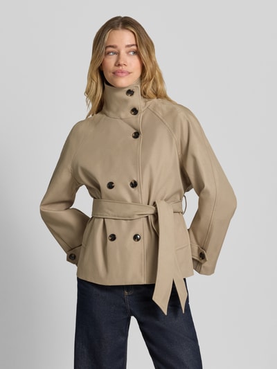 Kobiece model nosi trencz Trench Jacke mit Bindegürtel Modell 'Munet' od Neo Noir z bawełny, prosty krój, stójka, listwa guzikowa 2-rzędowa, wiązany pasek, patki przy mankietach, wpuszczane kieszenie; długość tyłu w rozmiarze 36: 62 cm, szerokość ramion: 50 cm, szerokość w rozmiarze 36: 47 cm, długość rękawów: 56 cm, jasnobeżowy kolor, gładka faktura, całkowity widok z przodu.