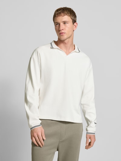 Männermodel trägt Strickpolo aus reiner Baumwolle Modell 'JORNIAGARA' von JACK & JONES in Weiß mit V-Ausschnitt, lockerer Passform, Kontrast-Umlegekragen, elastischen Ärmelabschlüssen und Label-Detail, Rückenlänge bei Größe M 64 cm, Ganzkörperansicht von vorne.