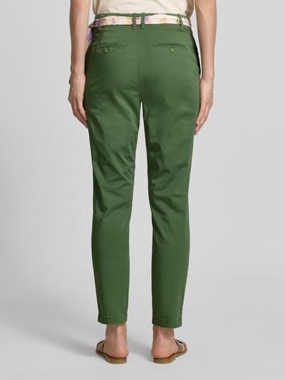 Kobieta nosi Slim Fit Chino mit Viskose-Anteil od Christian Berg Woman, zielone chinosy, mieszanka bawełny i wiskozy z dodatkiem wiskozy, krój slim fit, skrócony fason, odpinana materiałowa tasiemka, zapięcie na guzik i zamek błyskawiczny, widoczne tylne kieszenie, zdjęcie całej sylwetki z tyłu.