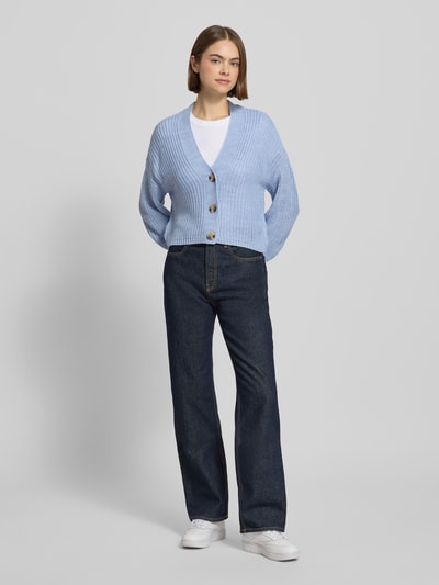 Frauenmodel trägt Regular Fit Strickjacke mit einreihiger Knopfleiste Modell 'CAROLSPRING' von Only, Optik: Unifarben in Strickoptik, cropped Länge, tiefer V-Ausschnitt, Langarm, gerippte Abschlüsse, Label-Stitching, Ganzkörperansicht von vorne.