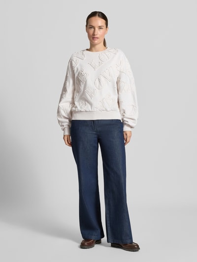 Modelka kobieca nosi białą, ozdobioną naszywką bluzę Oversized Sweatshirt aus reiner Baumwolle Model 'ASTERA' od DRYKORN, czysta bawełna, krój oversized, szerokie ramiona, napis, ściągacze, widok całej sylwetki z przodu.