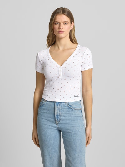 Vrouwenmodel draagt Cropped T-shirt met V-hals en knoopsluiting van Guess Jeans, materiaalmix met viscose, wit met logo, ruglengte maat S 43 cm. Ganzkörperansicht van voren.